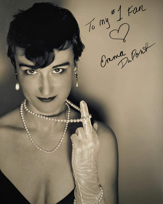 A headshot showing Myriam as Emma DuPont looking sultry at the viewer while holding up her middle finger, her pearl necklace draped from her finger to her neck. The headshot is signed in character by Emma to her number one fan.